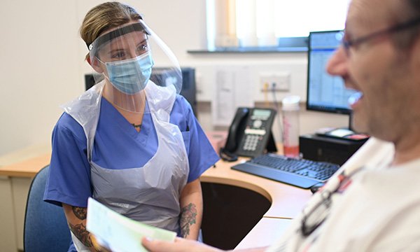 A general practice nurse wearing full PPE and talking to a patient during a consultation