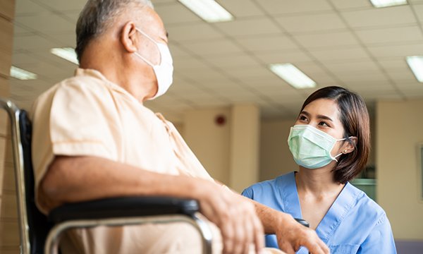 A masked nurse talking to a masked male patient. After backlogs due to COVID-19, people with cancer need to know nurses are there for them, to advise, to care and to listen
