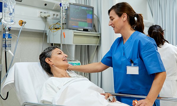 A nurse in uniform on the ward, standing talking to a patient in a hospital bed, indicating how mental health nursing skills can support the care of patients being treated for physical conditions