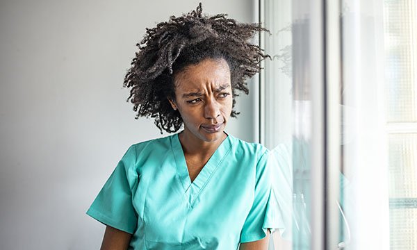 A nurse in green scrubs looks out a window looking pensive and anxious