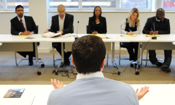 A man with his back to the camera raises his hands as he speaks to a panel of five people sitting opposite him