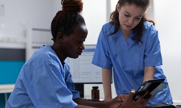 A seated nurse holds an electronic pad while a second nurse standing next to her looks at it