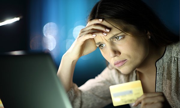 A young woman holds a hand to her brow and looks perplexed as she looks at a computer screen, holding a credit card in her other hand