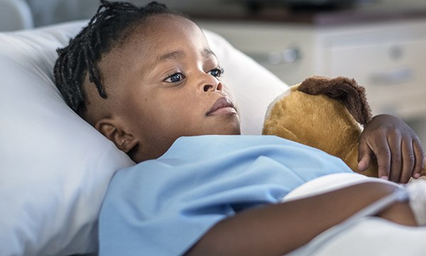 Picture shows a young black boy lying in a hospital bed, clutching a toy
