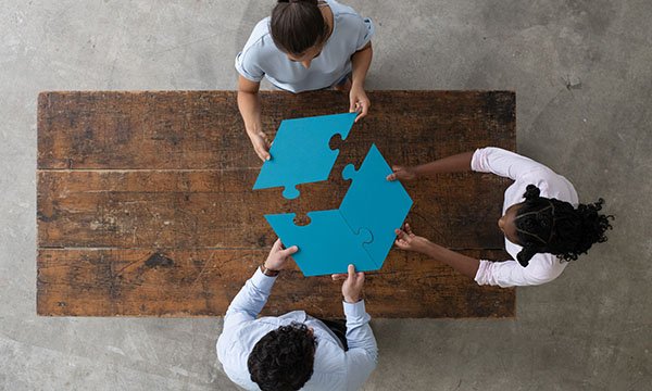 Three people sit at a table with jigsaw pieces that they are putting together, this illustrates how to make services fit correctly and work efficiently