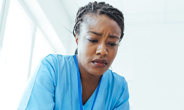 Nurse in scrubs tunic leans leans forward, frowning. She is alone and looks strained