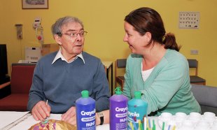 A nurse sits talking to a nursing home resident as he undertakes a craft activity