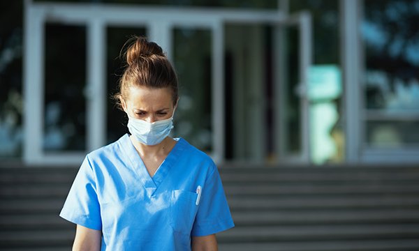 Nurse wearing a face mask walks away from a hospital building, looking downcast