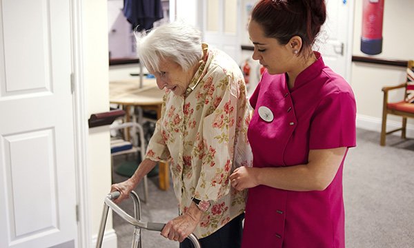 Picture shows a carer helping an older woman who is walking with the help of a zimmer frame
