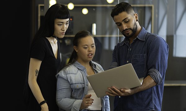 Two women and a man share and discuss ideas standing in a huddle around a laptop