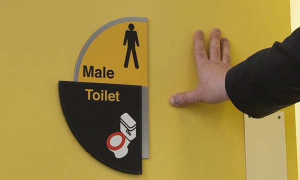 A hand pushes open the door of a male toilet which has dementia signage affixed to it