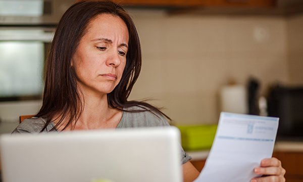 A woman sitting at a computer frowns as she holds up a bill